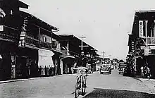 Man bicycling down street in old photo, with cars in background