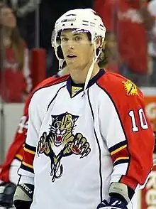 A Caucasian ice hockey player in his mid-twenties pictured from the waist up. Dressed in full equipment, he wears a white jersey with orange trim and a logo of a panther. He stands relaxed on the ice looking slightly to his left.