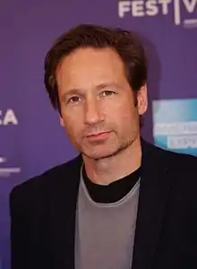 A man with dark brown hair is smirking and looking near the camera.