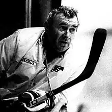 Black and white photo of a middle-aged man wearing a white jacket, hockey gloves, and holding a hockey stick