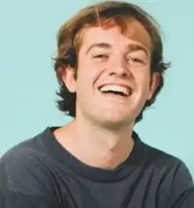 A Caucasian male with blonde hair, smiling at the camera against a bright blue background.