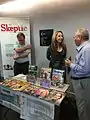 Deborah Hyde, editor of The Skeptic, standing behind the booth of the magazine, talking to attendees of the 16th European Skeptics Congress in London.