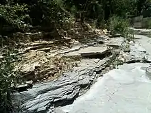 This photo shows a bluish layer of claystone below a greyish slab and a layer of yellow and brown above that. Where this more impervious rock forms the bottom of the creek, there is flowing water all year.