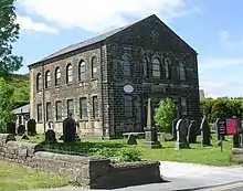 A plain dark stone church with two tiers of windows, the upper round-headed, and a small round window above the entrance in the gable end, in a graveyard and with countryside beyond