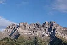 Vertical sawtooth-shaped rock walls overhanging a green slope lined with a few trees.