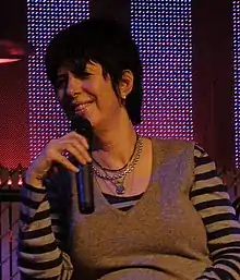 Diane Warren looking to the right and smiling while holding a microphone in her hand.