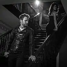 A black-and-white image of Bentley wearing a leather jacket on a staircase, looking at a woman wearing a dress.