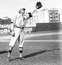 A man wearing a baseball uniform bearing the letter "P" on the chest leans slightly–his left and raises his left hand on which he is wearing a baseball glove.