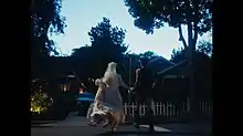 Two women (one in a wedding dress, the other in black commando clothing) hold hands as they run to a wedding getaway car. The setting shows a typical neighborhood at dusk hours.