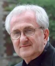 an older white man in a tight headshot. He is wearing glasses and a black priest's collar