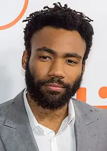 A closeup of a man smiling in front of a poster. He has a beard and black hair.