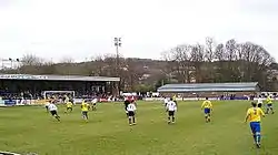 Two teams competing in a football match, one wearing white shirts and black shorts and the other yellow shirts and blue shorts.  A stand full of spectators and another low brick building are visible outside the playing area.