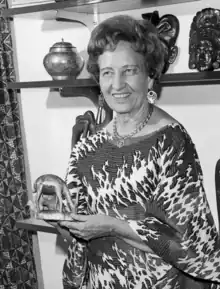 a woman dressed in African dress and jewelry holds a small sculpture of an African animal. Other artifacts line the shelves behind her.