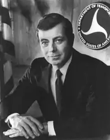 Black-and-white photo of a man wearing a suit sitting at a desk with his hands folded on it and the DOT logo and US flag behind him