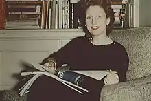 A middle-aged woman sits in a chair with a broad open smile looking at the camera with an open book on her lap and a bookcase of books in the background. She wears a full-body black dress, white pearl necklace, white pearl earrings and a gold bracelet.