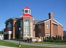 A large brick building of irregular polygonal shape rests amid a green lawn with ornamental shrubbery by a curving paved driveway. An ornamental lamp post rises above the driveway, and a United States flag rises on a pole near the building and opposite the lamp post. The building's bell tower rests on a two-story base fitted with elaborate arched windows.