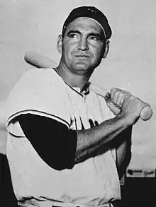 A man in a white baseball jersey and dark cap rests a bat on his shoulder.