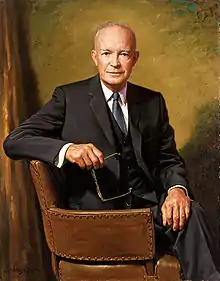 A balding man with gray hair, seated and holding his eyeglasses in his right hand. He is wearing a black suit, white shirt, and blue tie