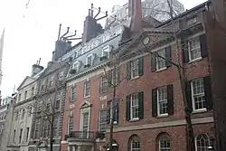 Four townhouses with two trees in front and ornate facades seen from across the street and to their right. They are all three stories high. The one on the far left is light tan, the one on the center left is brown brick, and the two right ones are regular red brick.