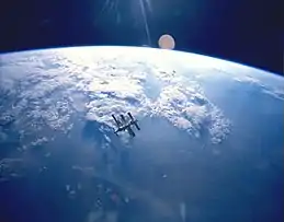 A cluster of modules and feathery solar arrays floats in the middle distance before an image of the Earth and the blackness of space above its horizon. Sunrays project from the top centre of the image.