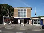 A brown-bricked building with a blue sign reading "EASTCOTE STATION" in white letters and people walking in front all under a blue sky with white clouds