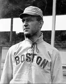 A man wearing a baseball cap and jersey with "Boston" written across the chest shown from the waist up looks–the left.