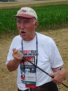 Man in a ball camp and white Tshirt holding a thin rod and standing in a field