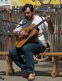 Eduard Drach wearing blue jeans and a white long-sleeved shirt, sitting on a bench, playing acoustic guitar and singing into a microphone