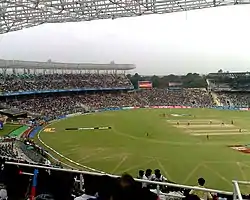 View from the top of a cricket ground during a cricket match, focusing on one side of the crowd; players are visible in the field.