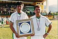 Two men in white football jerseys holdin a plaque.