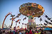 People line up in front of a moving chair swing ride full of riders. The sun is setting in the background.