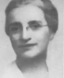 A smiling white woman with greying hair dressed back from her face, wearing glasses