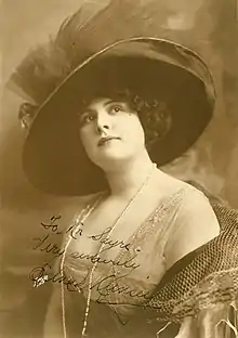 A young white woman with dark hair, wearing a large hat, a low-cut beaded gown, and pearls.