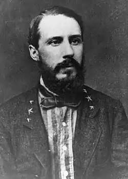 Photo shows a bearded man wearing a civilian coat over a striped shirt. The coat has three stars on each lapel.