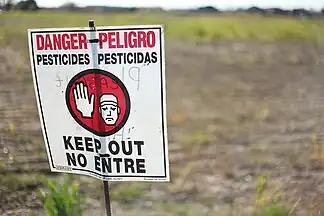 Eerie red and white caution sign signaling to not enter the designated area behind the sign. In red and black bold letters "DANGER--PELIGRO PESTICIDES--PESTICIDAS KEEP OUT NO ENTRE". An image of a cartoon man holding his hand up signaling a universal sign of stopping.