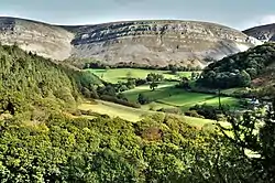 In the foreground is a wooded landscape, giving way to fields bounded by hedges, and low, round, bare hills in the backgound.