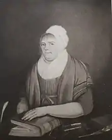 Portrait of Elizabeth "Betty" Beecroft by an unknown artist. Black and white picture of an oil painting. Betty is shown wearing a white cap, a dress and shawl. She has her arm resting on a book. Spectacles and writing equipment are on the desk in front of her.