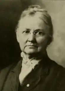 An older white woman, her grey hair in an updo, wearing glasses, a high-collared blouse, and a dark jacket