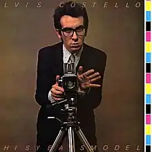 A young man wearing glasses and a suit-and-tie behind a camera on a tripod against a brown background