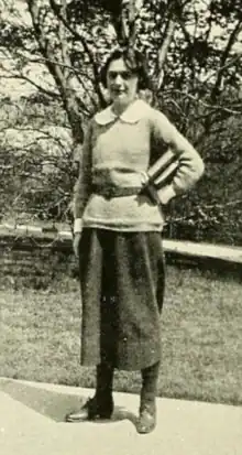 A young white woman with short dark hair, standing outdoors, wearing a peter-pan collar, a sweater belted at the waist, and a dark skirt; she has one hand on her hip