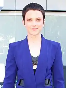 An upper body shot of the 22-year-old artist, who has close-cropped, dark hair with green-blue coloured eyes, a slight smile, and an upper left ear clasp. The artist wears a dark blue jacket with black trim at the pockets and is standing in front of a building with a row of windows behind.