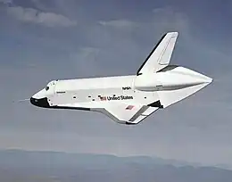 Enterprise glides to the left in this view, banked slightly towards the camera, showing "United States" on the port side and American flags on the port side and the port wing. Enterprise has a long probe sticking forward of the nose cone for calibration of instruments, and an aeroshell covering the engine area. Most of the backdrop is sky, but at the bottom, the mountainous horizon of California can be seen through haze.