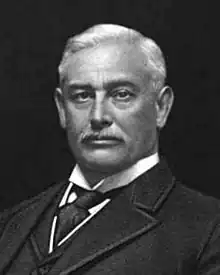 A black-and-white lithographic portrait of a middle-aged, mustached man from the shoulders up, wearing a suit, with a necktie