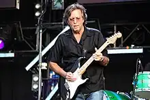 A man on a stage wearing blue jeans, a black shirt and eyeglasses. He is holding a black guitar, and part of a drum set and other audio and stage equipment can be seen in the background.