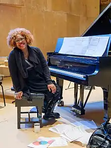 Errollyn Wallen on a chair, leaning to look towards the camera, at a piano.