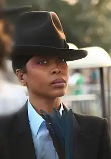 An image of a woman with a fedora and a suit holding blue feathers while staring forward with a neutral expression