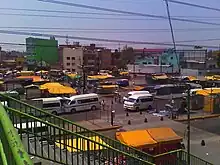 View of the bus terminal outside La Raza station Line 3. Several Nissan Caravan minibusses are parked. There are several retailers in the area.