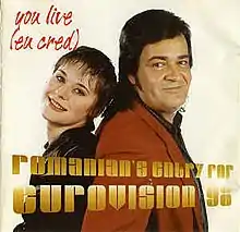 A shot of Olinescu and Adrian Romcescu standing side to side in front of a beige backdrop. Information on the song is superimposed on them in yellow and red letters.