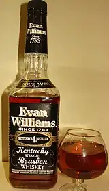 A color photograph of a bottle of bourbon to the left of a glass containing some of that bourbon