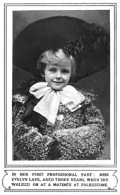 White female child with large hat and huge bow around her neck, smiling at the camera